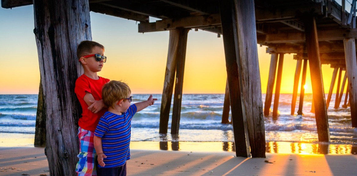 Two boys standing under the pier at sunrise | Williamson Realty Vacations Ocean Isle Beach Rentals